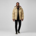 Gekleurde man gekleed in een zwarte broek en een wijd streetwear-jack met beige capuchon op een lichtgrijze achtergrond.