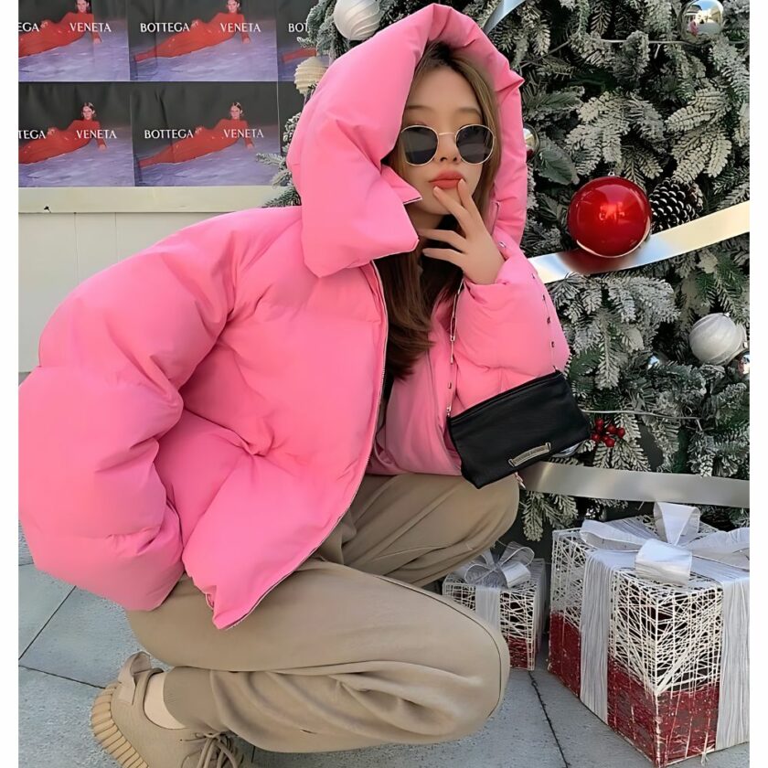 Vrouw hurkt met zonnebril voor een kerstboom, draagt een roze donsjack en capuchon over haar hoofd.