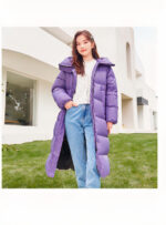 Jonge brunette vrouw met lang haar draagt een witte trui onder een lange, dikke, paarse jas met een open vrouwencapuchon en een hand in de zak van haar blauwe spijkerbroek. Er is groen en een wit gebouw met een erker op de achtergrond.