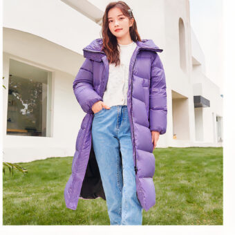 Jonge brunette vrouw met lang haar draagt een witte trui onder een lange, dikke, paarse jas met een open vrouwencapuchon en een hand in de zak van haar blauwe spijkerbroek. Er is groen en een wit gebouw met een erker op de achtergrond.