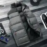 Grijs en zwart gewatteerd mouwloos donsjack met halfopen zakken met rits op donkergrijze strokenvloer. Aan de zijkanten zitten een zwarte pet, een zwarte trainer en een beetje een nummerplaat.