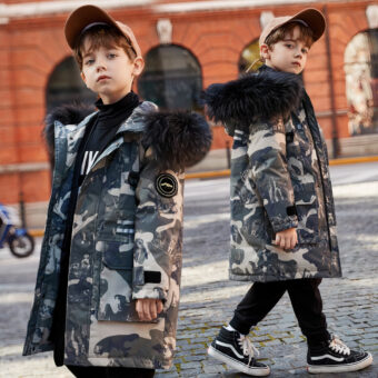 We zien een jonge stadsjongen met een beige pet, een zwarte spijkerbroek en zwarte sportschoenen, en een donsjack met militaire opdruk en een capuchon met zwart bont.