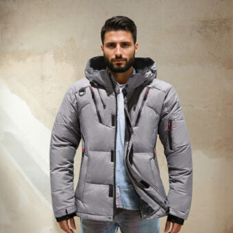 Bruinharige man met zijn rug naar een lichtbruine muur, hij draagt een spijkerbroek, een lichtblauw shirt en een grijs gewatteerd jack met capuchon.