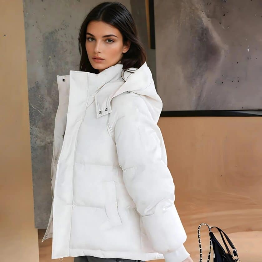 Bruinharige vrouw in profiel met een korte witte donsjas met capuchon, een handtas achter haar en een beige en grijze muur.