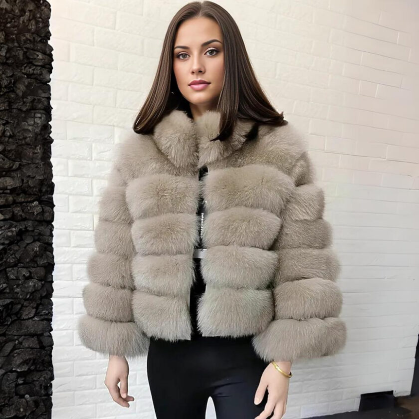 Kastanjeharige vrouw staat met haar rug naar een witte stenen muur, draagt een beige donsjack van imitatiebont en een zwarte broek.