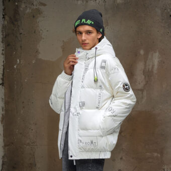 Jonge man met een grijze pet met groen opschrift, draagt een wit gewatteerd donsjack met opdruk op de capuchon en een zwarte broek. Zijn linkerhand in de zak van het donsjack, de andere houdt de bovenkant van het donsjack vast. In profiel met een bruine muur erachter.