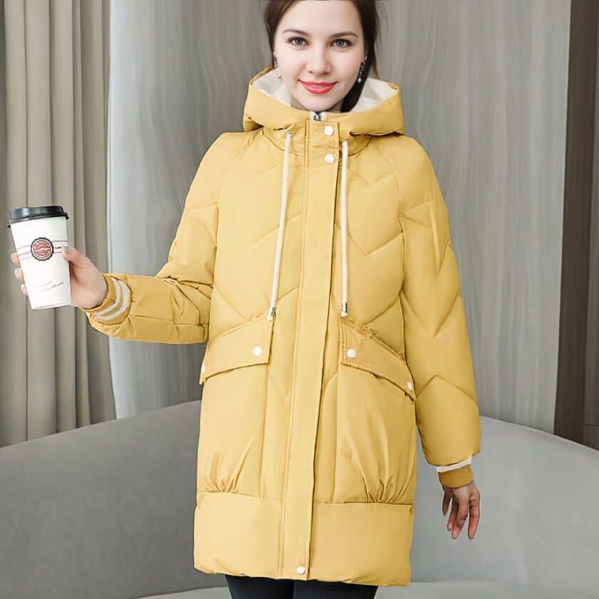 Een vrouw met een gele halflange jas met capuchon en koffie in haar hand, staat voor een grijze bank.