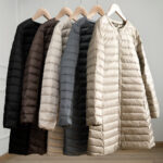 5 lichte en lange donsjassen in wit, grijs, zwart en bruin hingen op hangers.