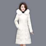 Bruine Aziatische vrouw draagt een lange, gewatteerde witte donsjas met bontkraag. Haar rechterhand in de zak van de donsjas. Grijze achtergrond.