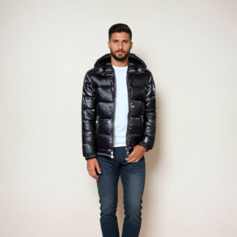 Een ongeschoren donkerharige man met een blauwe spijkerbroek, een wit T-shirt en een glanzend zwart gewatteerd jack met capuchon, die voor een beige muur staat.