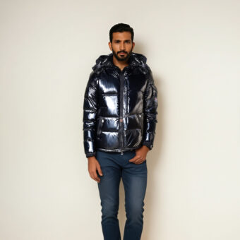 Bruinharige man staat tegen een beige muur, draagt een blauwe spijkerbroek en een glanzend zwart jack met capuchon.