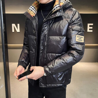 We zien een man in een elegante hotellobby, gekleed in het zwart met een glanzend zwart donsjack en een beige geruite voering op de capuchon.