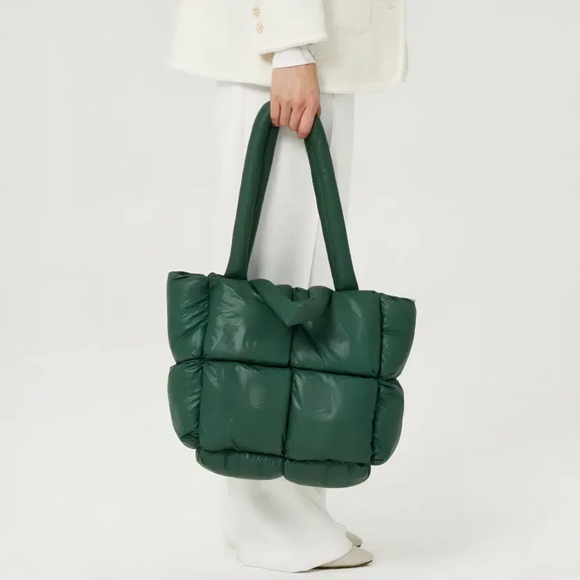 Een vrouwenbeen in profiel met witte broek op een witte achtergrond, met een grote groene gewatteerde plunjezak met een geïntegreerde schouderriem.