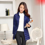 Vrouw draagt een lange, gewatteerde blauwe donsjas en houdt een tijdschrift vast, met een fauteuil en een lamp op de achtergrond.