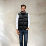 Donkerharige man in een donkerblauwe spijkerbroek, een witte top met lange mouwen en een zwart gewatteerd mouwloos jack, die met zijn rug naar een beige muur staat.