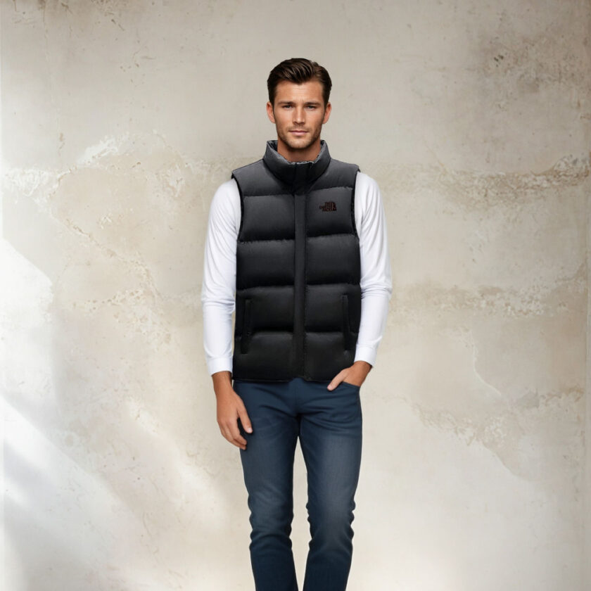 Donkerharige man in een donkerblauwe spijkerbroek, een witte top met lange mouwen en een zwart gewatteerd mouwloos jack, die met zijn rug naar een beige muur staat.