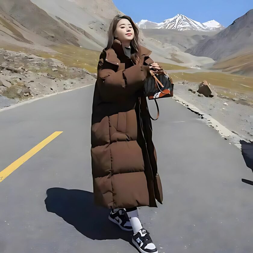 Vrouw met een lange bruine donsjas, staat op een weg midden in de bergen.