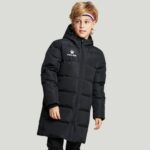 Een jonge blonde jongen draagt een lang gewatteerd katoenen sportjack voor kinderen en poseert voor een grijze achtergrond.