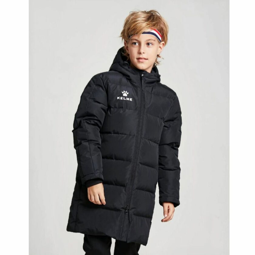 Een jongen tegen een grijze achtergrond met een lange zwarte gewatteerde donsjas met capuchon en een driekleurige hoofdband in de kleuren van de Franse vlag in zijn lichtbruine haar.