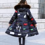 Bruinharige vrouw, staand van achteren met een beige stenen muur tegenover zich, haar hoofd naar rechts gedraaid. Ze draagt een zwarte broek en een lange, omkeerbare donsjas in marineblauw met witte, rode en groene motieven en letters. De capuchon heeft bont rond de rand.