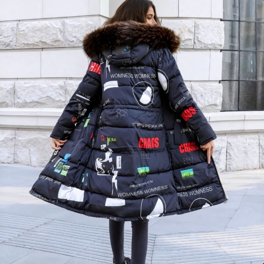 Bruinharige vrouw, staand van achteren met een beige stenen muur tegenover zich, haar hoofd naar rechts gedraaid. Ze draagt een zwarte broek en een lange, omkeerbare donsjas in marineblauw met witte, rode en groene motieven en letters. De capuchon heeft bont rond de rand.