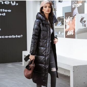 Staande vrouw met rechts op de achtergrond een muur met schrift en links een zwarte muur met wit schrift. Achter de vrouw staat een grijze stenen bank. Ze draagt een zwarte dlimbroek met een gat in de linkerknie, een beige pet, een lange zwarte capuchon en een handtas in haar rechterhand.