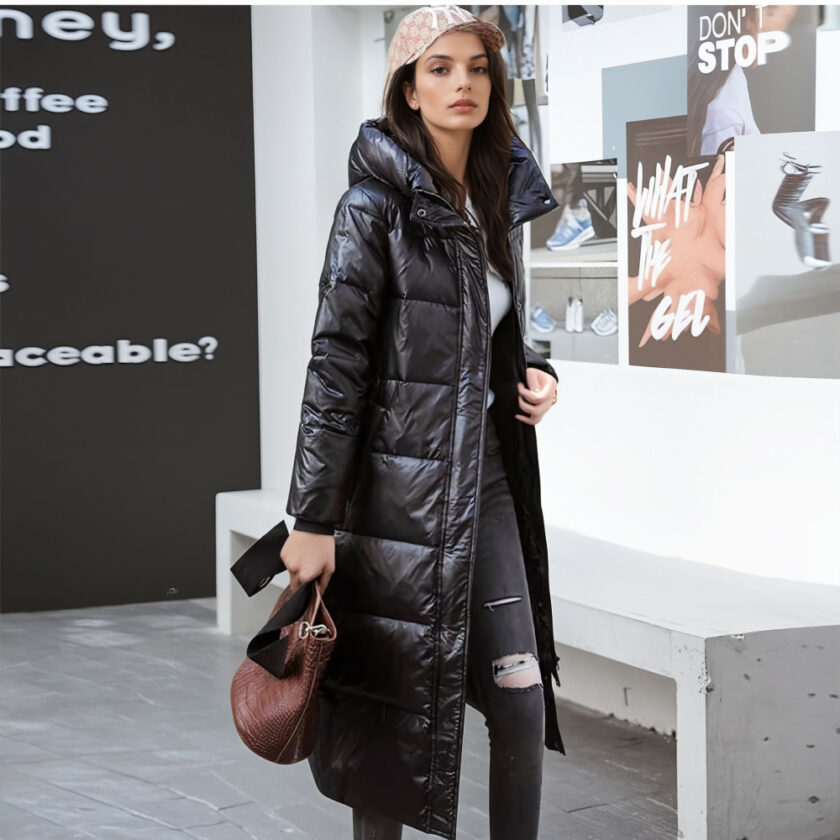 Staande vrouw met rechts op de achtergrond een muur met schrift en links een zwarte muur met wit schrift. Achter de vrouw staat een grijze stenen bank. Ze draagt een zwarte dlimbroek met een gat in de linkerknie, een beige pet, een lange zwarte capuchon en een handtas in haar rechterhand.