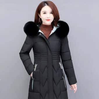 Aziatische vrouw met halflang bruin haar draagt een lange, gewatteerde, getailleerde zwarte donsjas met zwart bont op de capuchon. Ze houdt haar rechterhand in de zak van de donsjas. Grijze achtergrond.