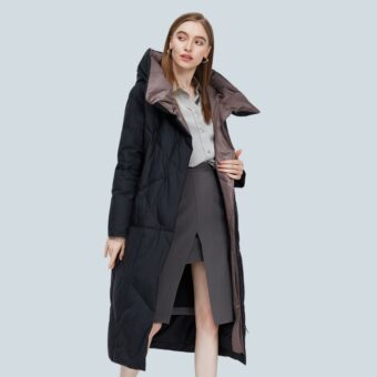 Een vrouw met lang, steil kastanjebruin haar draagt een lange, zwarte, open donsjas met een grote bruine kraag aan de binnenkant. Daaronder is ze gekleed in een lichtgrijs shirt en een donkergrijze kokerrok.