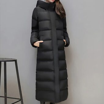 Donkerharige vrouw met een dikke, lange donsjas met capuchon in een grijze kamer met een kruk aan de linkerkant.