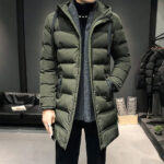 Man draagt een lange groene gewatteerde donsjas, met zwarte achtergrond en lichte parketvloer.