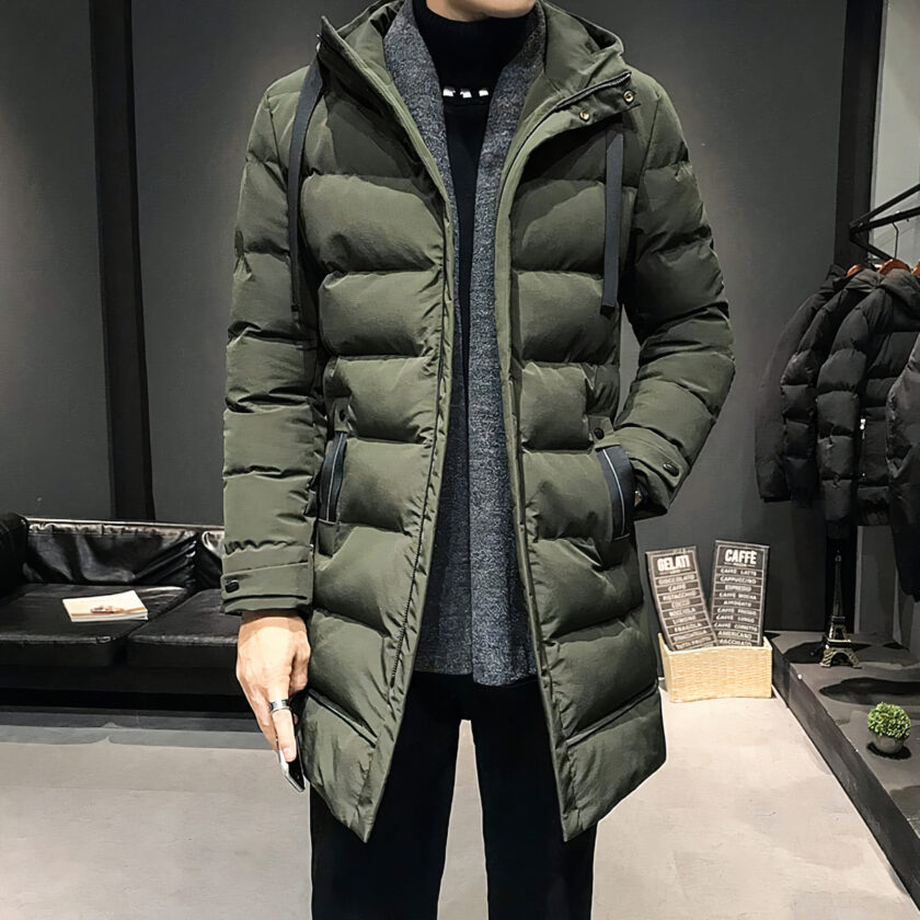 Man draagt een lange groene gewatteerde donsjas, met zwarte achtergrond en lichte parketvloer.