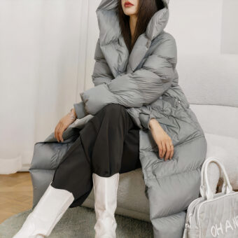 Vrouw zit op een bank en draagt een lange grijze donsjas met een zwarte broek en witte schoenen.