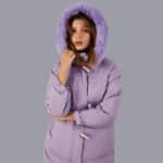 Vrouw draagt een paarse donsjas met een bontmuts over haar hoofd, op een grijze achtergrond.