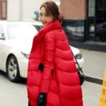 Foto van een vrouw in een lange rode donsjas die over straat loopt, met linksachter een witte auto en rechtsachter een gele bink.