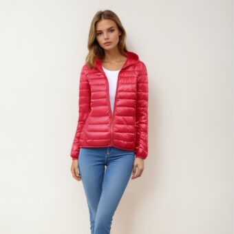 Blonde vrouw staat met haar rug tegen een beige muur, draagt een blauwe skinny jeans, een wit topje en daarover een licht, warm rood donsjack dat voor een derde dichtgeritst is, waardoor het witte topje zichtbaar wordt.