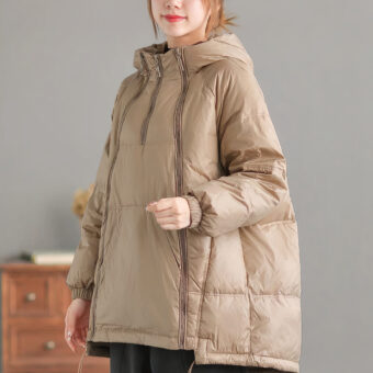 We zien een vrouw in een kamer met een houten ladekast en een vrij donkergrijze muur. Ze draagt een zwangerschapsjas: een beige jas die tijdens de zwangerschap gedragen wordt met een kangoeroezak om de baby in te dragen als hij geboren is.