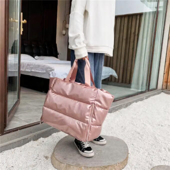 Grote gewatteerde tas in metallic roze wordt door een persoon in de hand gedragen. De persoon draagt een wit sweatshirt en een blauwe spijkerbroek en staat buiten op een dunne boomstam op een witte grindvloer. Er is een slaapkamer te zien, met een bed op de achtergrond achter een erker.