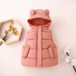 Een roze mouwloos donsjack met capuchon en oren voor een klein meisje, gepresenteerd op een pastelroze achtergrond naast katoenen bloemen en een boeket gele bloemen in een frame.