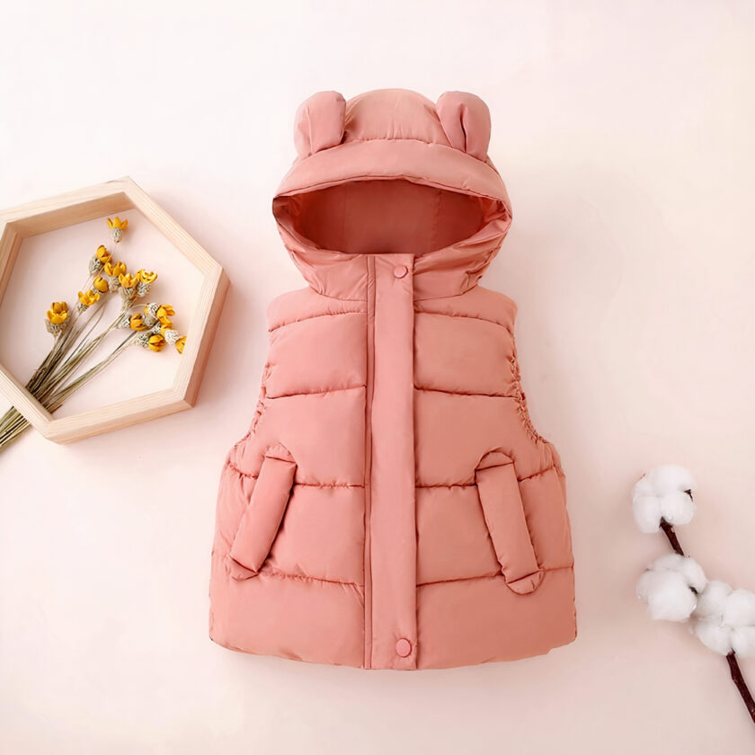 Een roze mouwloos donsjack met capuchon en oren voor een klein meisje, gepresenteerd op een pastelroze achtergrond naast katoenen bloemen en een boeket gele bloemen in een frame.