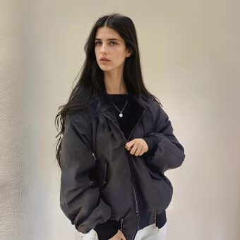 Een slanke, brunette vrouw met lang, los haar, die een zwart omkeerbaar jack draagt en met haar rug naar een beige muur staat.