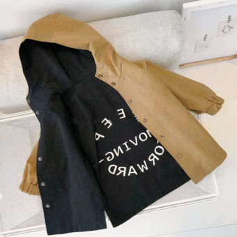 De foto is van bovenaf genomen en toont een omkeerbare windjackjas voor kinderen: de ene kant is zwart, de andere camel, met witte letters op de achterkant.