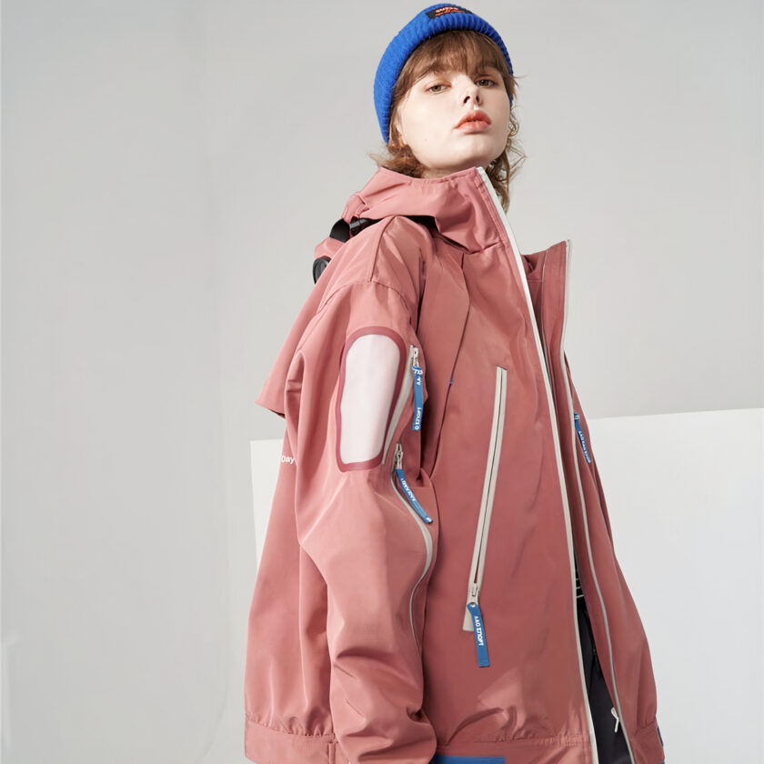 Vrouw met blauwe muts die een roze halflange ski-jas voor vrouwen draagt op een grijze achtergrond.