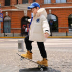 We zien een tiener met een blauwe pet over straat skateboarden, met een witte donsjas met capuchon op die er erg warm uitziet.