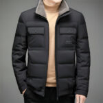 Kort, mat zwart jack met fleece kraag en twee zakken met drukknopen op de borst, halfopen gedragen door een man met een beige trui eronder.