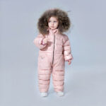 Klein meisje dat een roze donsjack met volledige bescherming draagt met de capuchon over haar hoofd voor een grijze achtergrond.