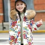 Jong meisje op straat draagt een witte donsjas met capuchon voor tieners De donsjas heeft een capuchon van beige bont Over de hele jas zijn gekleurde motieven gedrukt Het jonge meisje draagt een marineblauwe pet.