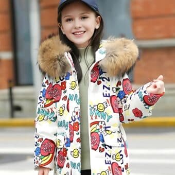 Jong meisje op straat draagt een witte donsjas met capuchon voor tieners De donsjas heeft een capuchon van beige bont Over de hele jas zijn gekleurde motieven gedrukt Het jonge meisje draagt een marineblauwe pet.