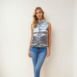 Blonde vrouw staat tegen een beige muur en draagt een blauwe skinny jeans en een zilverkleurig mouwloos donsjack.