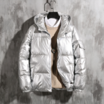 Foto van een zilveren donsjack van een man op een hanger voor een gewaxte betonnen muur, gedragen met een beige trui en een wit overhemd.